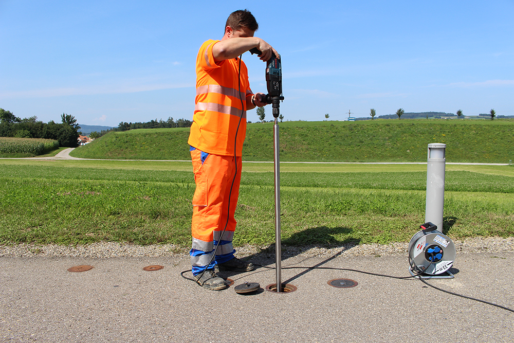 Photo d'un collaborateur en combinaison orange au bord d'une route en gravier à côté d'une prairie verte, montrant comment le remplacement d'EBG peut être effectué sans tranchée.